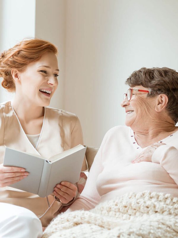 happy-nurse-reading-a-funny-book-to-her-elderly-ZPKCERN.jpg